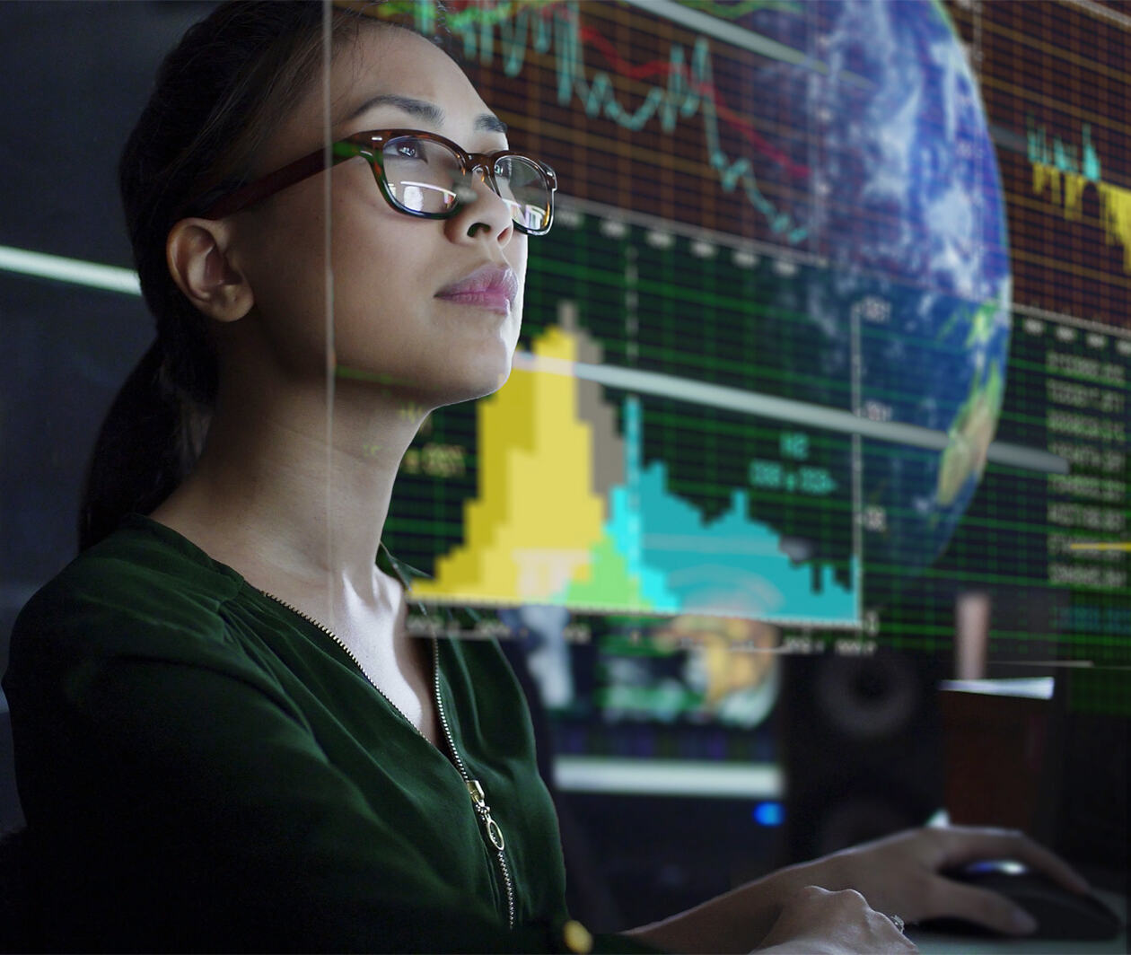 A young Asian woman looking at the global & environmental data projected on a see through display whilst seated in a dark office