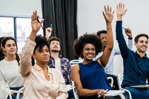 Adult students in a classroom, smiling and raising their hands.