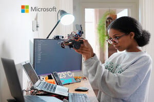 A young girl holding up a small robot she has built. Her two laptops, mobile phone, monitor and display panel are all open and active.